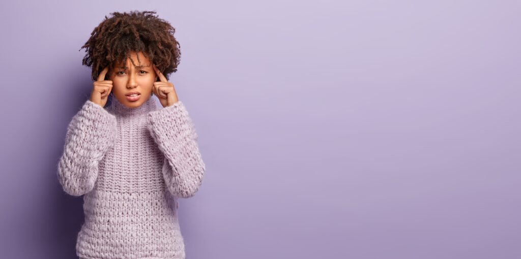 Young woman with curly hair wearing a cozy lavender sweater, looking stressed and pressing her fingers to her temples, standing against a plain purple background.