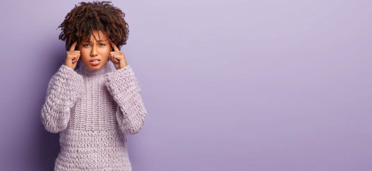 Young woman with curly hair wearing a cozy lavender sweater, looking stressed and pressing her fingers to her temples, standing against a plain purple background.